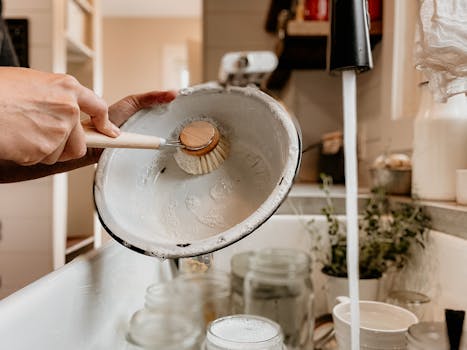 Une main nettoie un plat en métal avec une brosse, sous un robinet qui coule, dans une cuisine minimaliste.
